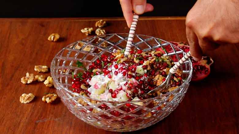 Two hands are seen holding salad spoons and tossing a salad with pomegranates, cucumbers, walnuts, and yogurt in a large cut-glass bowl. California walnuts kernels and an open pomegranate lie on the wooden counter in the background - Konkani Cucumber and Pomegranate Salad Recipe